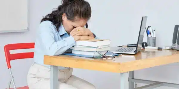 Exhausted teacher leaning on stack on students books on her desk to rest after long stressful day