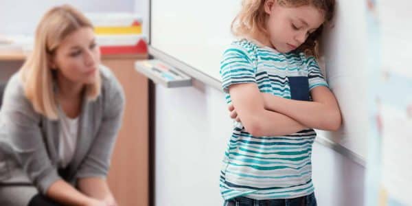 Offended child stands with his arms folded in the classroom, his back to the teacher